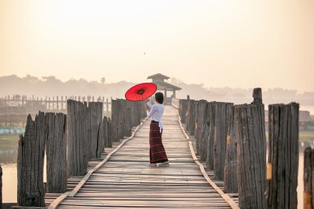 668965135 - ผู้หญิงกำลังถือร่มสีแดง. เดินบนสะพานอูเบ็ง